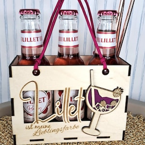 May include: A wooden carrier holds three bottles of Lillet, with red and white labels, and a burgundy handle. The carrier has a cut-out design with the word "Lillet" and a cocktail glass illustration. Two copper-colored straws are included.