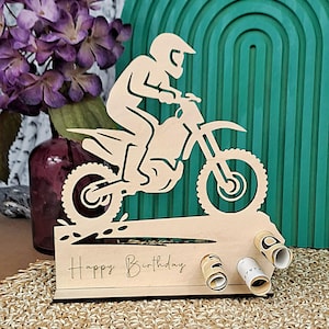 Op de afbeelding: Een houten verjaardagskaart met een motorcrosser op een motorfiets. De kaart heeft de tekst "Happy Birthday" en bevat kleine opgerolde rollen. De achtergrond omvat een vaas met paarse bloemen en een groene boogvormige decoratie.