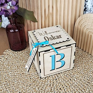 Puede incluir: Un bloque de madera con la letra "B" azul y el nombre "Oskar" en la parte superior. El bloque está atado con una cinta azul y tiene una etiqueta con texto en alemán. Está sobre una superficie tejida.