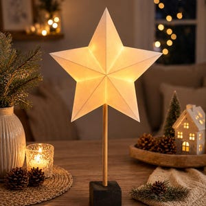 May include: A five-pointed star lamp with a white paper shade emitting a warm glow. The star is mounted on a slender, light-colored wooden rod attached to a solid, dark gray square base, creating a freestanding decorative lighting fixture.