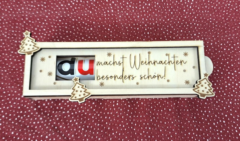 Pode incluir: Uma caixa retangular de madeira com tampa deslizante, decorada com pequenas &aacute;rvores de Natal de madeira e a frase "du machst Weihnachten besonders sch&ouml;n!". Cont&eacute;m uma barra de chocolate Kinder.