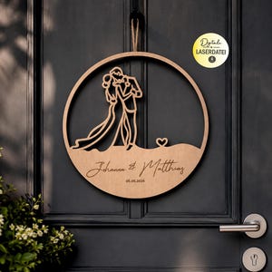 Könnte beinhalten: Ein hölzerner Hochzeitskranz mit kreisförmigem Design. Der Kranz zeigt eine Silhouette eines tanzenden Paares, ein Herz und die Namen "Johanna & Matthias" mit dem Datum "06.06.2026". Der Kranz hängt an einer dunkelgrauen Tür.