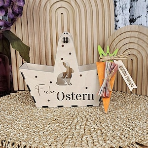May include: A wooden Easter basket with a bunny silhouette and the words "Frohe Ostern." A carrot-shaped gift tag with "Bunny Money" is attached. The basket is on a woven mat, with a wooden arch in the background.