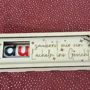Pode incluir: Uma caixa de madeira com um painel deslizante, apresentando a palavra "du" em vermelho e preto. A caixa &eacute; decorada com recortes de fadas e a frase alem&atilde; "zauberst mir ein L&auml;cheln ins Gesicht".