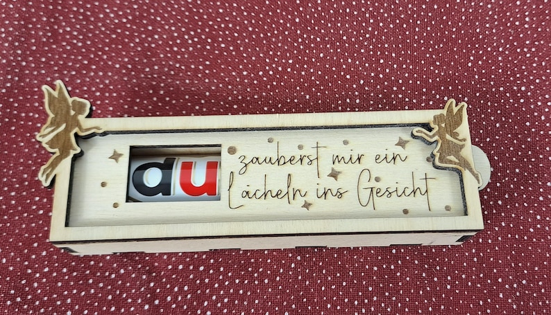 Pode incluir: Uma caixa de madeira com um painel deslizante, apresentando a palavra "du" em vermelho e preto. A caixa &eacute; decorada com recortes de fadas e a frase alem&atilde; "zauberst mir ein L&auml;cheln ins Gesicht".