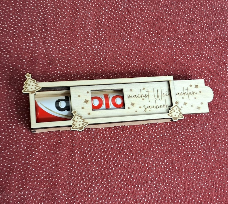 Pode incluir: Um suporte de madeira para Kinder Surpresa com um design deslizante. O suporte apresenta um design de &aacute;rvore de Natal e as palavras "macht Weihnachten zauberhaft". O ovo Kinder Surpresa &eacute; vis&iacute;vel no interior.