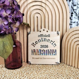 May include: A wooden retirement sign with the text "offiziell Rentnerin 2026 MARTINA DU HAST ES DIR VERDIENT!" is displayed. The sign is light brown and has a decorative border. Purple flowers are in a dark red vase.