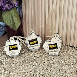 Puede incluir: Tres etiquetas de regalo de madera con forma de flor, corazón y flor con la palabra "Riesen" en cada etiqueta. Cada etiqueta tiene una frase alemana diferente. Las etiquetas están atadas con hilo natural.