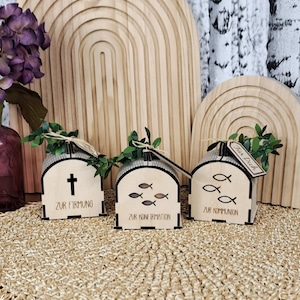May include: Three wooden gift boxes with arched roofs, each adorned with a different symbol: a cross, fish, and a tag. The boxes have the words "Zur Firmung", "Zur Konfirmation", and "Zur Kommunion" printed on them. Greenery and tags are attached.