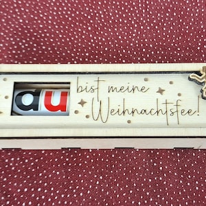 Pode incluir: Caixa de madeira com um painel deslizante, com o texto "du bist meine Weihnachtsfee!" e uma barra de chocolate Kinder Bueno. A caixa &eacute; decorada com recortes de fadas e estrelas. O fundo &eacute; um tecido vermelho com bolinhas.