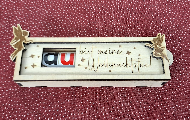 Pode incluir: Caixa de madeira com um painel deslizante, com o texto "du bist meine Weihnachtsfee!" e uma barra de chocolate Kinder Bueno. A caixa &eacute; decorada com recortes de fadas e estrelas. O fundo &eacute; um tecido vermelho com bolinhas.