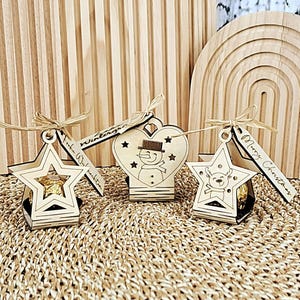 Könnte beinhalten: Drei Weihnachtsornamente aus Holz in Stern- und Herzform. Jedes Ornament hat ein kleines Geschenk im Inneren und ist mit einem festlichen Design verziert. Die Ornamente sind mit Bindfaden gebunden und tragen Anhänger mit der Aufschrift "Merry Christmas" und "Frohe Weihnachten".