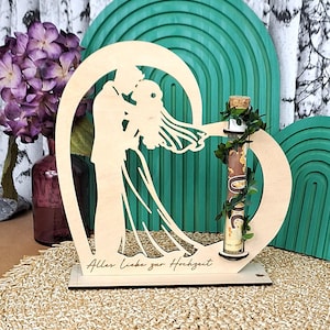 Könnte beinhalten: Ein hölzernes Hochzeitsandenken mit einer Braut- und Bräutigam-Silhouette in einem herzförmigen Rahmen. Ein Glasröhrchen mit Korken ist mit Grün geschmückt. Auf dem Sockel steht "Alles Liebe zur Hochzeit."