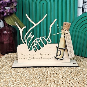 Peut inclure: Décoration de mariage en bois avec deux mains jointes, avec l'inscription "Hand in Hand ein Leben lang!". Une petite bouteille en verre avec un bouchon et une étiquette "Alles Liebe zur Hochzeit" est attachée.