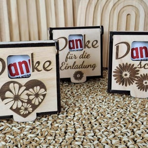 Könnte beinhalten: Drei Geschenkboxen aus hellem Holz mit dunkelbraunen Rändern. Jede Box hat einen Ausschnitt für einen kleinen Schokoriegel und das Wort "Danke" in Schreibschrift. Eine Box hat ein Herzdesign, eine andere sagt "für die Einladung" und die dritte "schön".