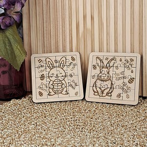 May include: Two wooden Easter jigsaw puzzles. Each puzzle features a bunny design and the words "Frohe Ostern". The puzzles are made of light-coloured wood and are square-shaped.