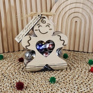 Könnte beinhalten: Ein Geschenk in Form eines Lebkuchenmannes aus Holz mit einem herzförmigen Ausschnitt, gefüllt mit verpackten Süßigkeiten. Das Etikett lautet: „Warm Christmas wishes for you and your family“. Das Geschenk ist in einem transparenten Cellophanbeutel verpackt.