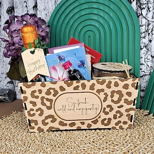 May include: A wooden gift basket with a leopard print pattern and the German text 'Sei frech, wild und einzigartig!' engraved on the front. It contains a small bottle with an orange foil top, a 'Happy Birthday!' tag, a floral card, and a candle.