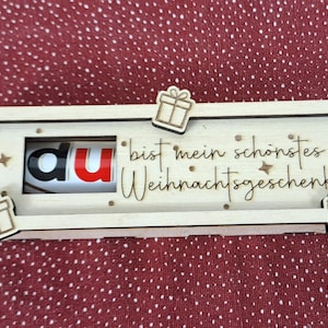 Pode incluir: Uma caixa de presente de madeira com tema natalino com as palavras "du bist mein sch&ouml;nstes Weihnachtsgeschenk" e uma barra de chocolate Kinder Bueno dentro. A caixa &eacute; decorada com pequenos recortes de caixas de presente e estrelas.