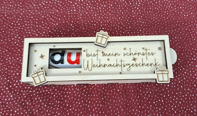 Pode incluir: Uma caixa de presente de madeira com tema natalino com as palavras "du bist mein sch&ouml;nstes Weihnachtsgeschenk" e uma barra de chocolate Kinder Bueno dentro. A caixa &eacute; decorada com pequenos recortes de caixas de presente e estrelas.