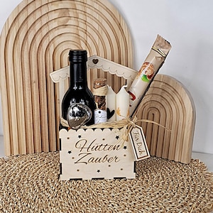 Könnte beinhalten: Ein hölzerner Geschenkkorb mit einer Weinflasche, Kerzen und einem Schokoriegel. Der Korb trägt die Aufschrift "Hütten Zauber". Das Geschenkset ist mit Bindfaden gebunden und enthält ein Etikett mit der Aufschrift "Für Dich".