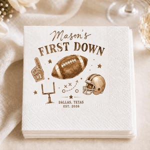 Op de afbeelding: Een vierkante witte papieren servet met bruine bedrukte afbeeldingen van een football, een helm, een doelpaal en een schuimvinger. De tekst luidt Mason's FIRST DOWN, DALLAS, TEXAS, EST. 2026. Het ontwerp bevat kleine sterren en spelschema's.