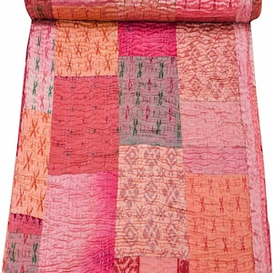 Può includere: Una trapunta patchwork nei toni del rosa, corallo e rosso. La trapunta è composta da vari quadrati di tessuto con diversi motivi e trame, tra cui punti a croce e disegni a righe. La trapunta è arrotolata nella parte superiore.