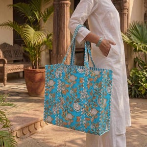 K&ouml;nnte beinhalten: Eine gro&szlig;e, rechteckige Tragetasche mit einem lebendigen Blumenmuster in T&uuml;rkis, Orange und Wei&szlig;. Die Tasche hat stabile Griffe und ein passendes gemustertes Innenfutter. Die Tasche ist aus einem gesteppten Stoff gefertigt.