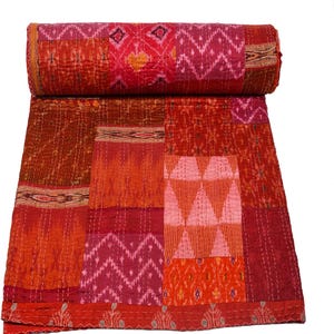 Peut inclure: Une courtepointe patchwork roulée aux tons de rouge, orange et rose. La courtepointe présente divers motifs et textures géométriques, notamment des chevrons et des triangles. Les bords sont finis avec une bordure décorative.