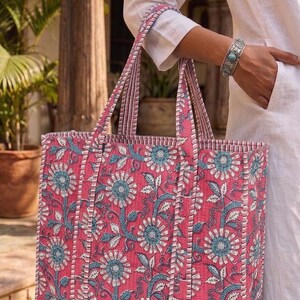 K&ouml;nnte beinhalten: Eine gro&szlig;e Tragetasche mit einem Blumenmuster in Rosa, Blau und Wei&szlig;. Die Tasche hat stabile Griffe und eine rechteckige Form. Das florale Design zeigt stilisierte Blumen und Bl&auml;tter. Die Tasche wird von einer Person in wei&szlig;er Kleidung gehalten.