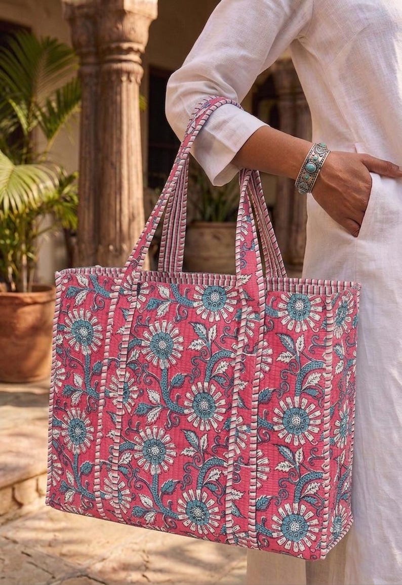 K&ouml;nnte beinhalten: Eine gro&szlig;e Tragetasche mit einem Blumenmuster in Rosa, Blau und Wei&szlig;. Die Tasche hat stabile Griffe und eine rechteckige Form. Das florale Design zeigt stilisierte Blumen und Bl&auml;tter. Die Tasche wird von einer Person in wei&szlig;er Kleidung gehalten.