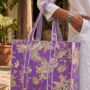 K&ouml;nnte beinhalten: Eine gro&szlig;e Tragetasche mit einem floralen Muster in Lila- und Beiget&ouml;nen. Die Tasche hat eine rechteckige Form mit stabilen Griffen und einem Streifenmuster an den Seiten. Die Tasche wird von einer Person gehalten, die ein wei&szlig;es Hemd tr&auml;gt.