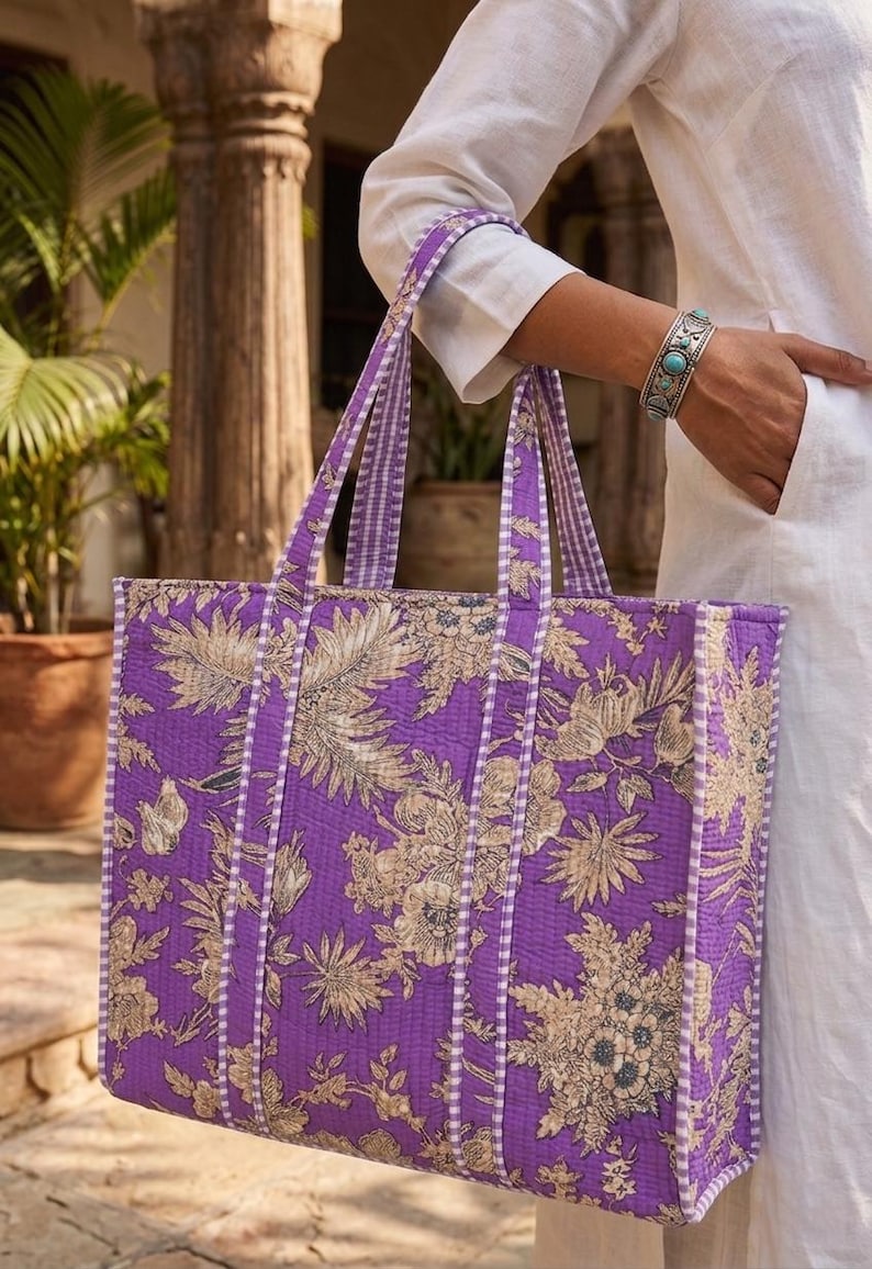 K&ouml;nnte beinhalten: Eine gro&szlig;e Tragetasche mit einem floralen Muster in Lila- und Beiget&ouml;nen. Die Tasche hat eine rechteckige Form mit stabilen Griffen und einem Streifenmuster an den Seiten. Die Tasche wird von einer Person gehalten, die ein wei&szlig;es Hemd tr&auml;gt.