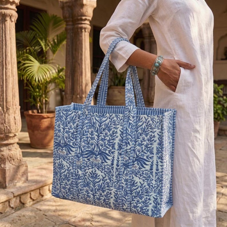 K&ouml;nnte beinhalten: Eine gro&szlig;e Tragetasche mit blauem und wei&szlig;em Blumenmuster. Die Tasche hat stabile Griffe und eine rechteckige Form. Das Design zeigt ein sich wiederholendes Muster aus stilisierten Bl&auml;ttern und Blumen. Die Tasche wird von einer Person in wei&szlig;er Kleidung gehalten.
