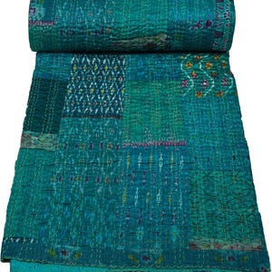 Op de afbeelding: Een turquoise en blauwgroene patchworkquilt met een opgerold bovenstuk. De quilt heeft verschillende patronen en zichtbare stiksels. Het kleurenpalet omvat tinten turquoise, blauwgroen en hints van paars en geel.
