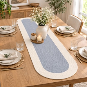 May include: An oval-shaped table runner featuring a braided texture with a central light blue panel bordered by a wide cream-coloured edge. The runner is placed on a wooden table set with white plates, woven placemats, and clear glassware.