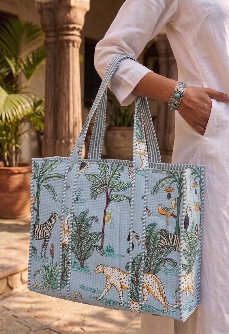 K&ouml;nnte beinhalten: Hellblaue Tragetasche mit Dschungel-Motiv, das Zebras, Leoparden und Palmen zeigt. Die Tasche hat gestreifte Griffe und ein gestepptes Design. Die Tasche wird von einer Person getragen, die ein wei&szlig;es Hemd und ein silbernes Armband tr&auml;gt.