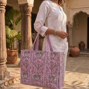 K&ouml;nnte beinhalten: Eine gro&szlig;e, gesteppte Tragetasche mit einem Blumenmuster in Rosa-, Gr&uuml;n- und Lilat&ouml;nen. Die Tasche hat lange Henkel und eine rechteckige Form. Der Hintergrund zeigt eine Person in einem wei&szlig;en Outfit.