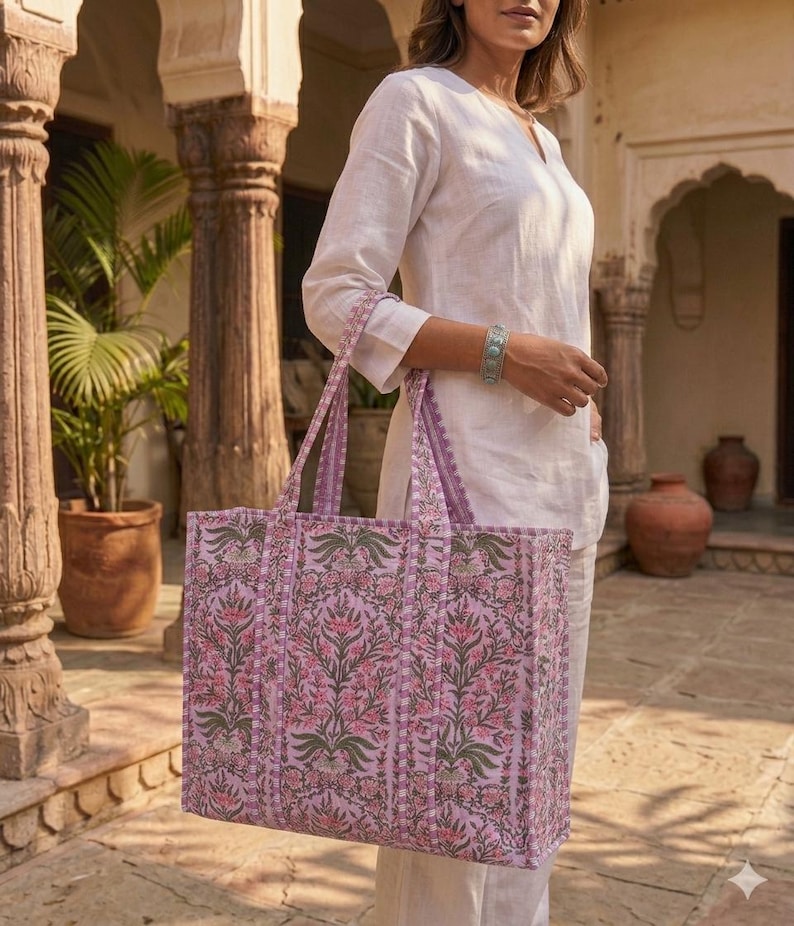 K&ouml;nnte beinhalten: Eine gro&szlig;e, gesteppte Tragetasche mit einem Blumenmuster in Rosa-, Gr&uuml;n- und Lilat&ouml;nen. Die Tasche hat lange Henkel und eine rechteckige Form. Der Hintergrund zeigt eine Person in einem wei&szlig;en Outfit.