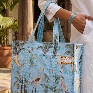 K&ouml;nnte beinhalten: Hellblaue Tragetasche mit einem sich wiederholenden Muster aus Leoparden, V&ouml;geln und tropischen Pflanzen. Die Tasche hat blau gestreifte Griffe und Verzierungen. Die Tasche ist aus einem gesteppten Stoff gefertigt.