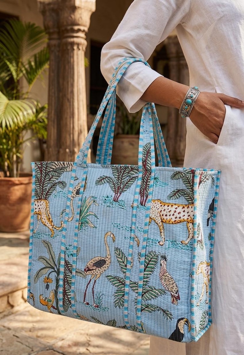K&ouml;nnte beinhalten: Hellblaue Tragetasche mit einem sich wiederholenden Muster aus Leoparden, V&ouml;geln und tropischen Pflanzen. Die Tasche hat blau gestreifte Griffe und Verzierungen. Die Tasche ist aus einem gesteppten Stoff gefertigt.