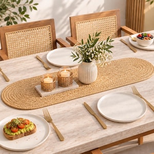Può includere: Un tavolo da pranzo in legno chiaro apparecchiato con piatti bianchi, posate dorate, un runner da tavola ovale intrecciato, un vaso bianco a coste con vegetazione e due candele in vetro in supporti intrecciati, circondato da sedie in legno.
