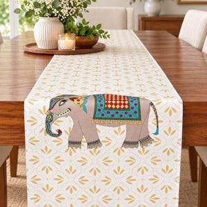 Peut inclure: Un chemin de table blanc avec un motif floral jaune répétitif et une illustration centrale d'un éléphant gris orné d'une couverture colorée, d'une coiffe décorative et de motifs complexes dans des tons de rouge, bleu et jaune.