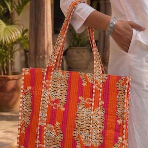K&ouml;nnte beinhalten: Eine gesteppte Tragetasche mit einem leuchtenden Tigerstreifenmuster in Orange und Pink. Die Tasche hat stabile Griffe und eine rechteckige Form. Das Design zeigt sich wiederholende Tigerillustrationen. Die Tasche wird von einer Person in wei&szlig;er Kleidung gehalten.