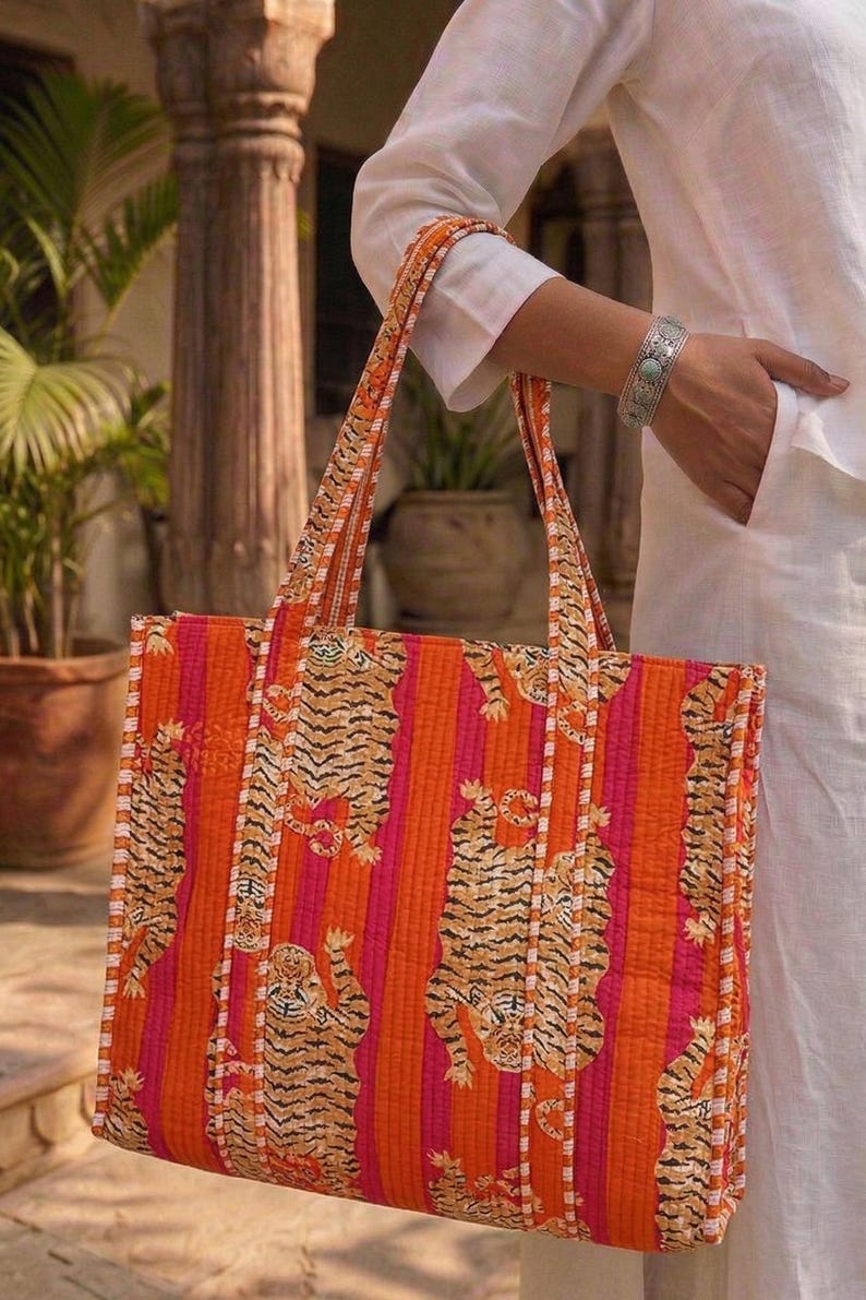 K&ouml;nnte beinhalten: Eine gesteppte Tragetasche mit einem leuchtenden Tigerstreifenmuster in Orange und Pink. Die Tasche hat stabile Griffe und eine rechteckige Form. Das Design zeigt sich wiederholende Tigerillustrationen. Die Tasche wird von einer Person in wei&szlig;er Kleidung gehalten.