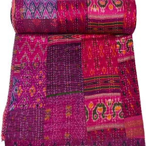 May include: A rolled-up patchwork quilt in shades of pink, magenta, and purple. The quilt features a variety of patterned fabric squares, including ikat and floral designs, stitched together with white thread. The edges are finished with a pink border.
