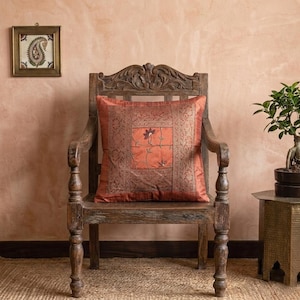 May include: An ornate, wooden armchair with a decorative backrest holds a square, rust-colored pillow. The pillow features a central square with a floral design and an intricate border. A small framed artwork hangs on the wall.