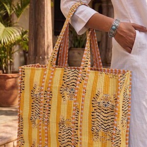 K&ouml;nnte beinhalten: Eine gro&szlig;e Tragetasche mit einem Tigerstreifenmuster in Gelb und Orange. Die Tasche ist aus einem gesteppten Stoff gefertigt und hat zwei Griffe. Der Hintergrund ist ein verschwommenes Bild einer Person, die die Tasche h&auml;lt.