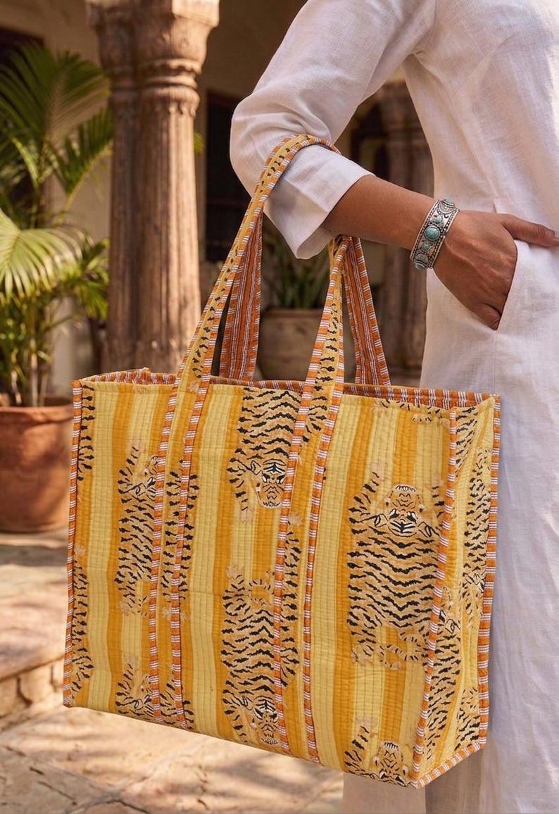 K&ouml;nnte beinhalten: Eine gro&szlig;e Tragetasche mit einem Tigerstreifenmuster in Gelb und Orange. Die Tasche ist aus einem gesteppten Stoff gefertigt und hat zwei Griffe. Der Hintergrund ist ein verschwommenes Bild einer Person, die die Tasche h&auml;lt.