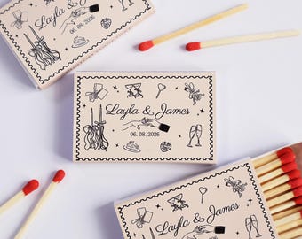 May include: Three rectangular matchboxes with a light beige cover featuring black line art of hands, champagne flutes, candles, a cake, and a strawberry. The text reads Layla & James and 06. 08. 2026. Several wooden matches with red tips are scattered around.
